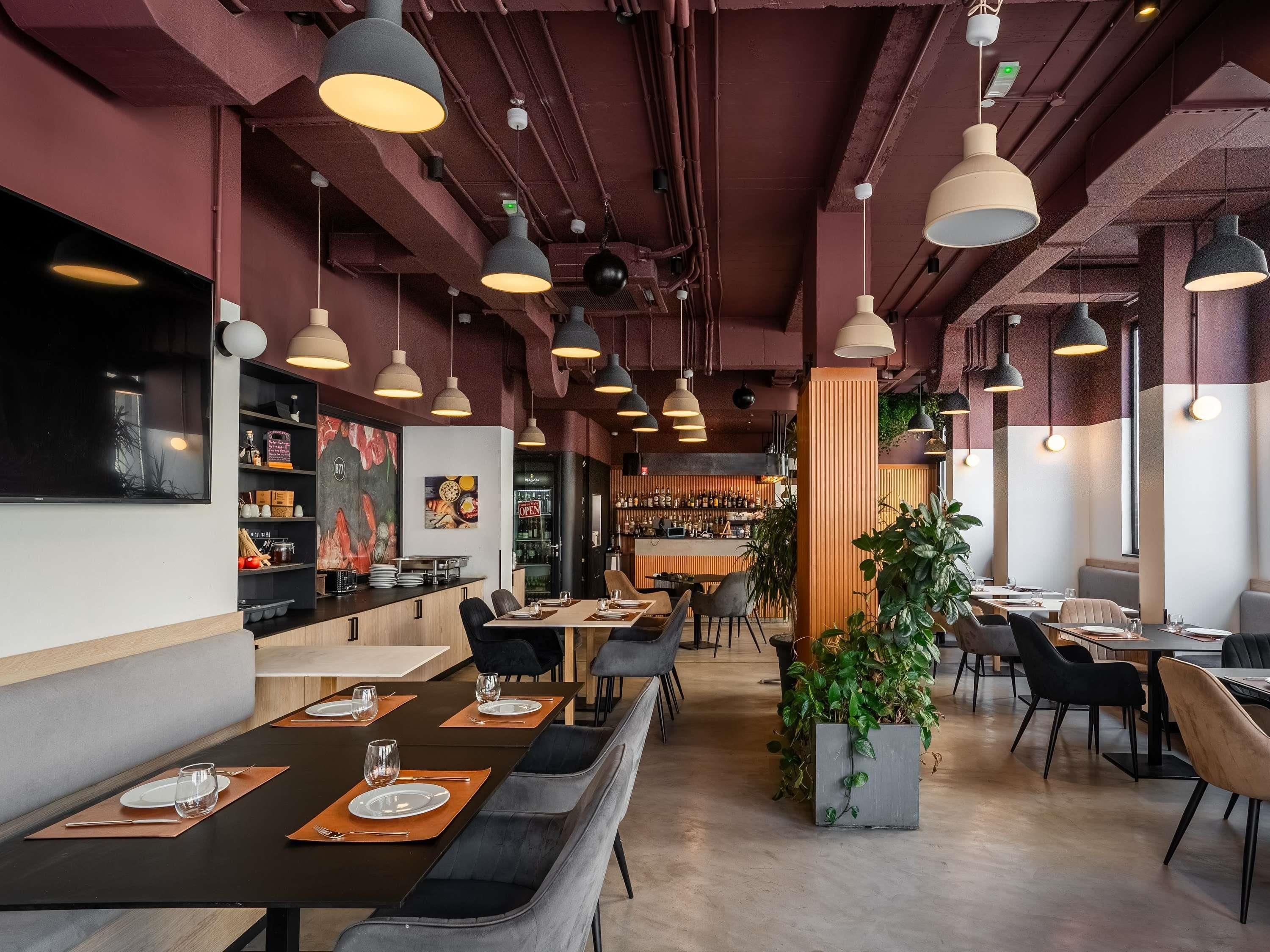 A restaurant interior with multiple dining tables, chairs, pendant lighting, a bar area, and a large potted plant.
