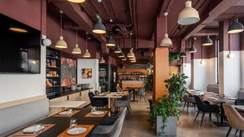 A restaurant interior with multiple dining tables, chairs, pendant lighting, a bar area, and a large potted plant.