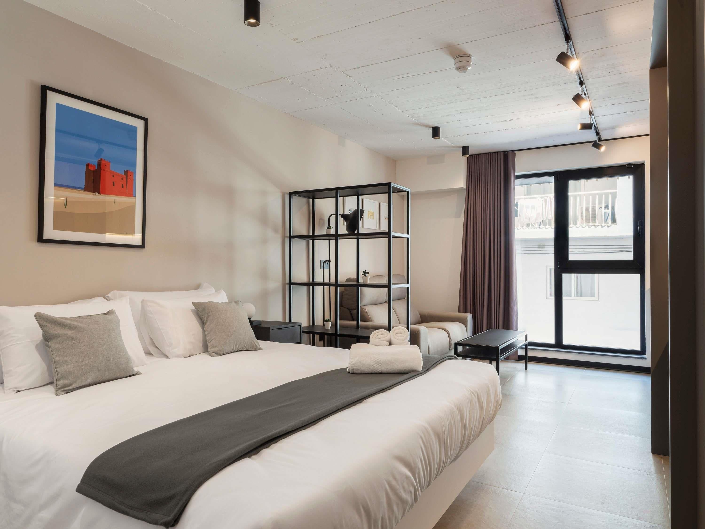 A modern hotel room with a large bed, a bookshelf, a desk, and a view of the outside.