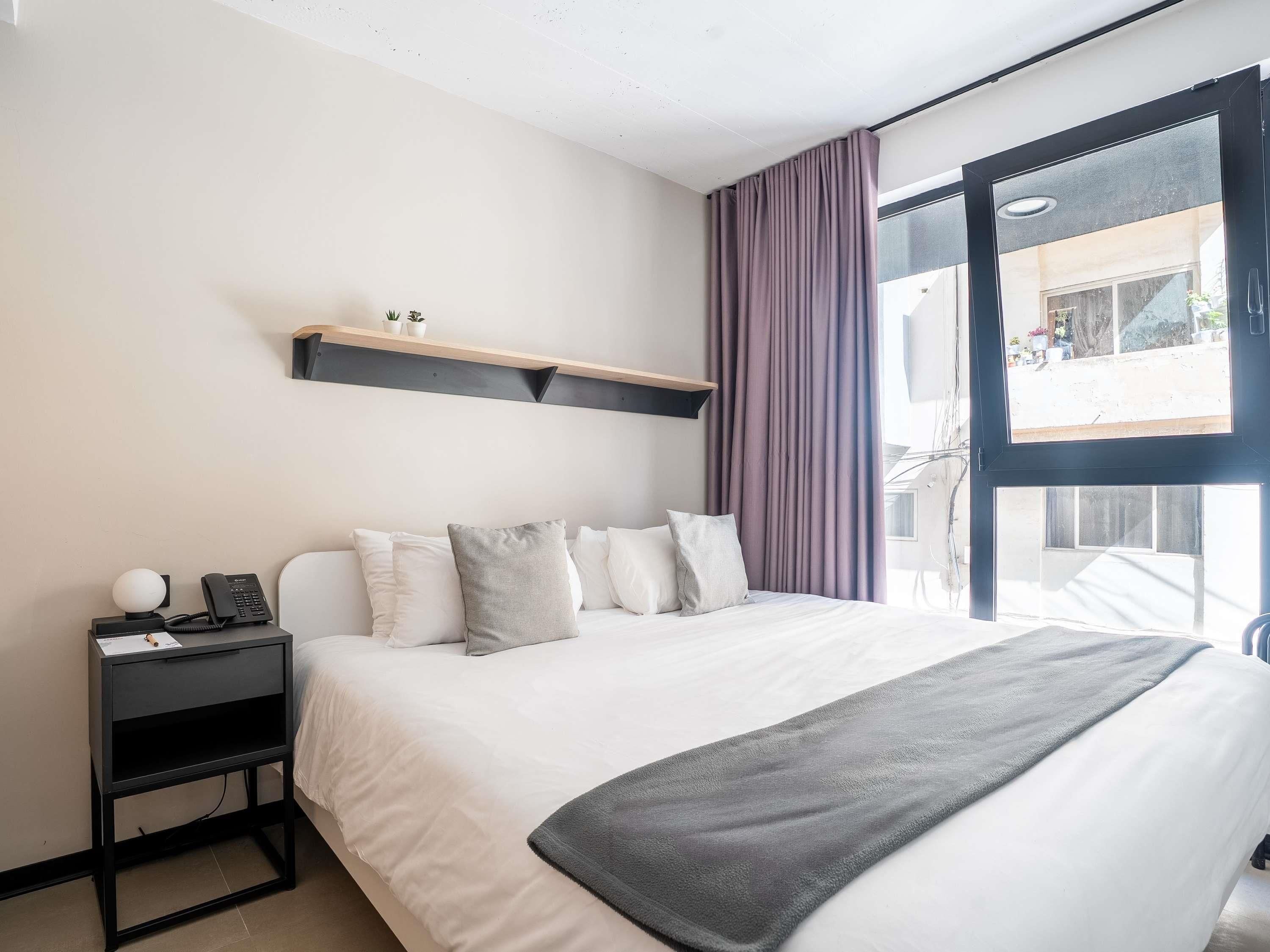 A modern hotel room with a large bed, a bedside table with a phone, a small shelf with decorative items, and a view of the outside through a large window.