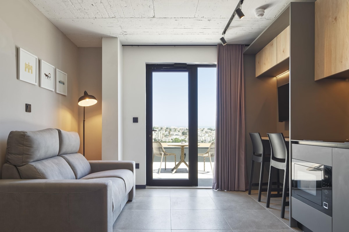 A modern living room with a sofa, a dining area with chairs, a kitchen with a microwave, and a view of the city through a glass door.