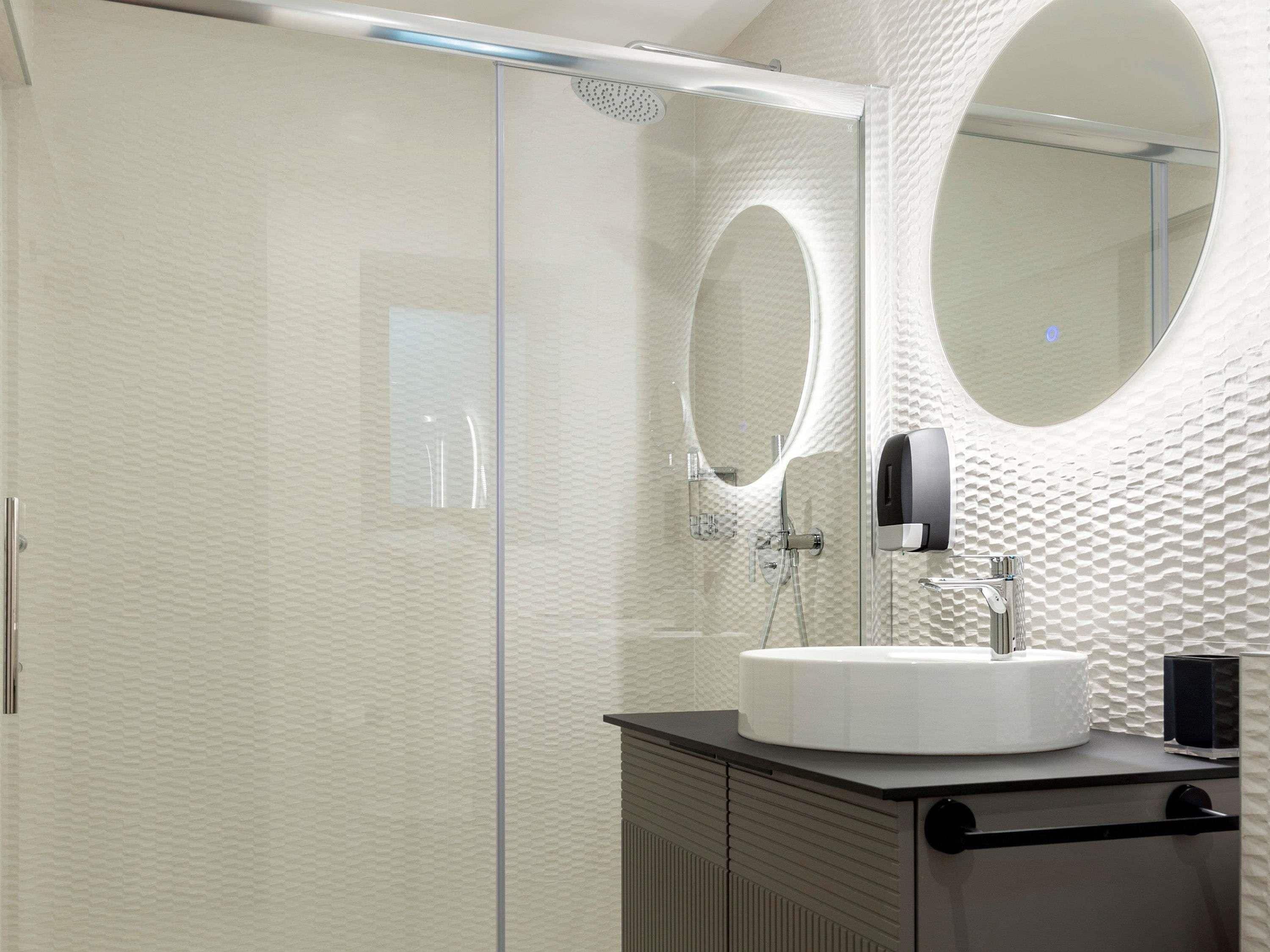 Un baño moderno con ducha de vidrio, espejo redondo y lavamanos blanco.