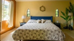 A bedroom with a bed, two nightstands, a lamp, a plant, and a window with a wreath. - Posada James (Xilitla)