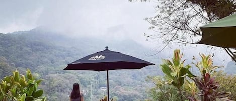 Una mujer está de pie junto a una mesa y sillas, con un paraguas negro, contemplando un paisaje montañoso.