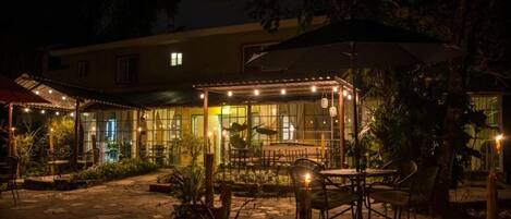 An outdoor patio area with tables and chairs, surrounded by plants and a covered seating area.