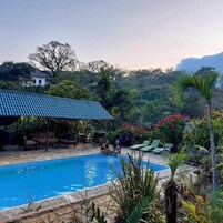 A swimming pool with lounge chairs and a covered seating area, surrounded by tropical vegetation and a mountainous backdrop.