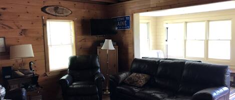 L’intérieur d’un chalet accueillant, avec un canapé en cuir, un fauteuil en bois, un téléviseur et une pancarte sur laquelle on peut lire : « VENEZ EN MAINE ».