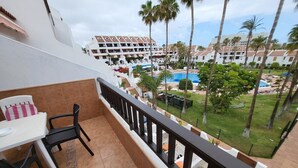 A balcony with a table and chairs, overlooking a pool area with palm trees and a hotel building in the background. - Parque Santiago II - 1 Bed (Playa de Las Americas)