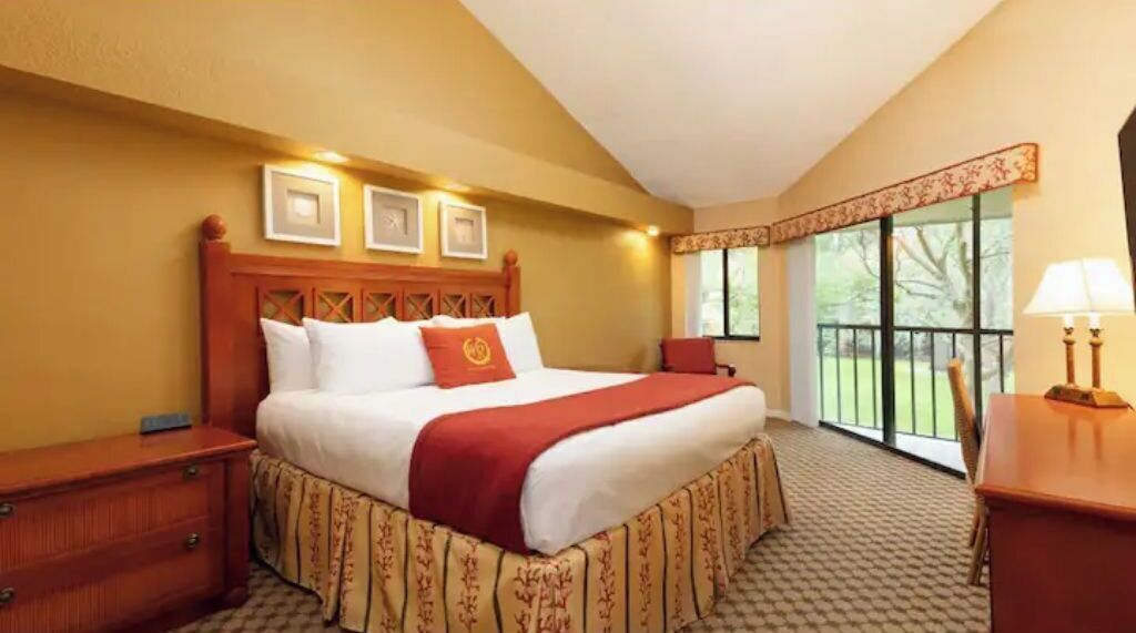 A hotel room with a large bed, a wooden headboard, a nightstand, a desk with a lamp, and a window with a view of greenery.