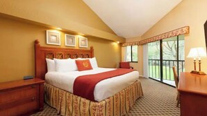 A hotel room with a large bed, a wooden headboard, a nightstand, a desk with a lamp, and a window with a view of greenery.