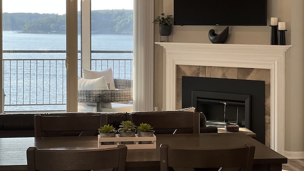 A dining area with a view of a lake, a fireplace, and a mounted television. - Pristine-Top Floor & Amazing Main Channel Views! Prime Location. (Lake Ozark)