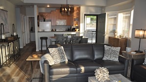 A modern living room with a black leather sofa, wooden coffee table, and a kitchen in the background. - Pristine-Top Floor & Amazing Main Channel Views! Prime Location. (Lake Ozark)