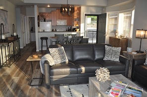 A modern living room with a black leather sofa, wooden coffee table, and a kitchen in the background.
