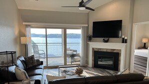 A living room with a fireplace, a large TV, and a balcony with a view of the water. - Pristine-Top Floor & Amazing Main Channel Views! Prime Location. (Lake Ozark)