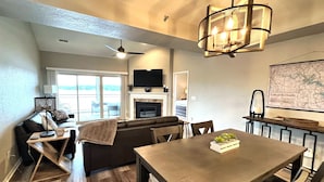 A modern living room with a fireplace, a dining area with a table and chairs, and a large window overlooking a deck. - Pristine-Top Floor & Amazing Main Channel Views! Prime Location. (Lake Ozark)
