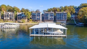 A floating structure with a covered deck, surrounded by residential buildings and a body of water. - Pristine-Top Floor & Amazing Main Channel Views! Prime Location. (Lake Ozark)