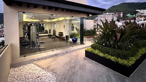 A modern gym with exercise equipment, glass walls, and a view of a cityscape. - Pavilion Paradise - Steps away from everything! (Puerto Vallarta)