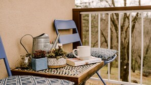 A wooden table with a metal watering can, a white mug, a book titled 'Have No Shame' by Melissa Foster, and patterned cushions.