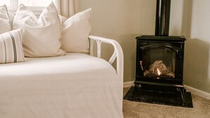 A bed with white bedding and pillows, a white metal frame, and a black wood-burning stove with a fire burning inside.