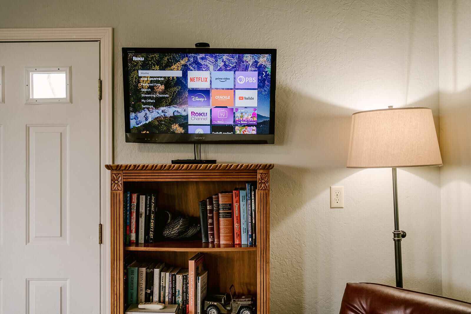 Ein flachbildfernseher an der Wand, der eine Streaming-Dienstanwendung anzeigt. Darunter befindet sich ein Holzregal mit Büchern und einer Fernbedienung. Eine Lampe ist rechts neben dem Regal platziert.