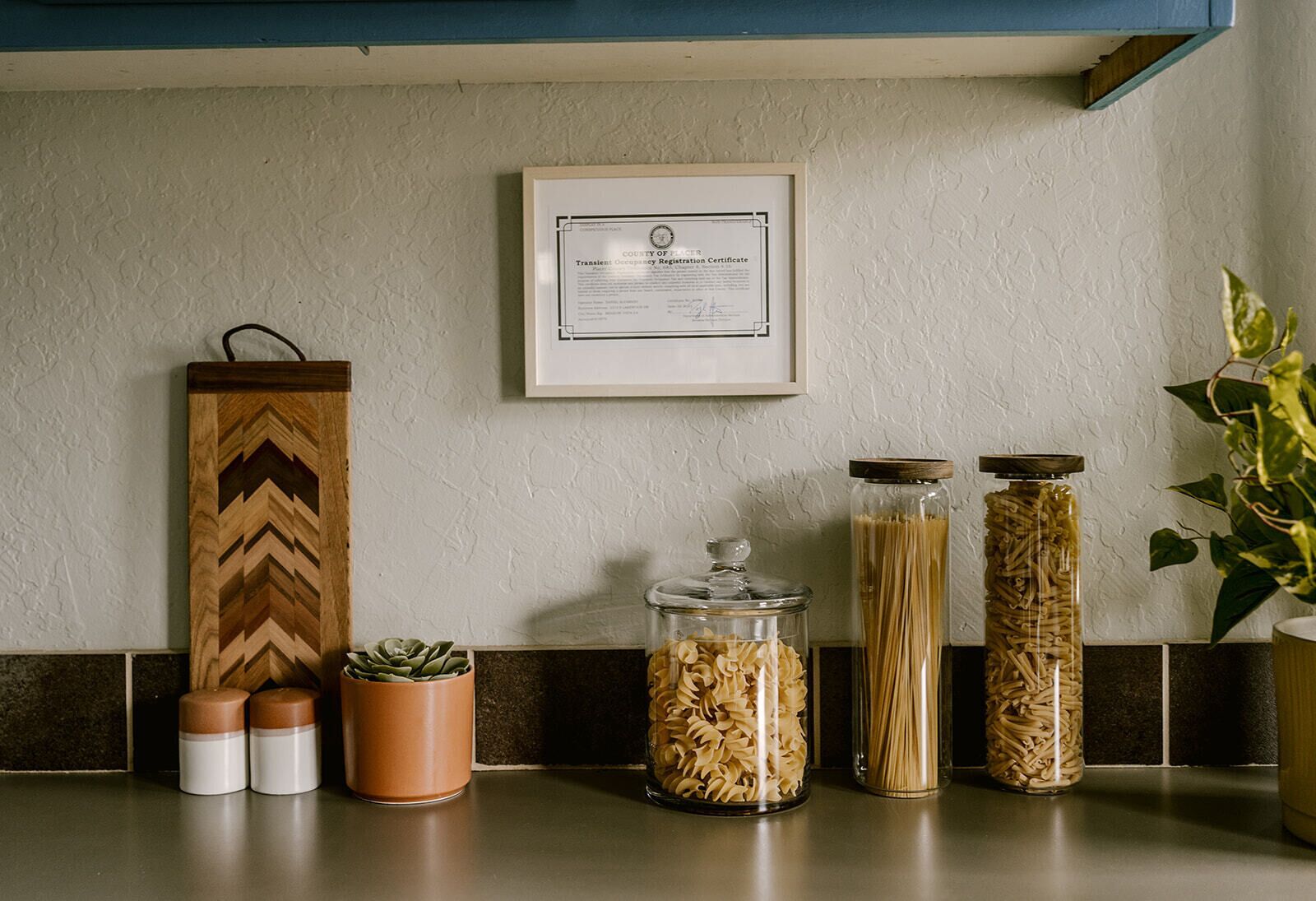 A kitchen counter with jars of pasta, a certificate on the wall, and a wooden decorative piece.