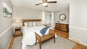 Um quarto com uma cama, ventilador de teto, cômoda, espelho e janela com cortinas.