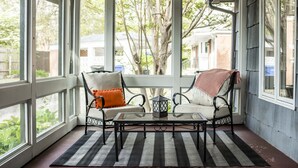 Varanda coberta com duas cadeiras, uma mesa e vista para uma árvore e casas ao fundo.