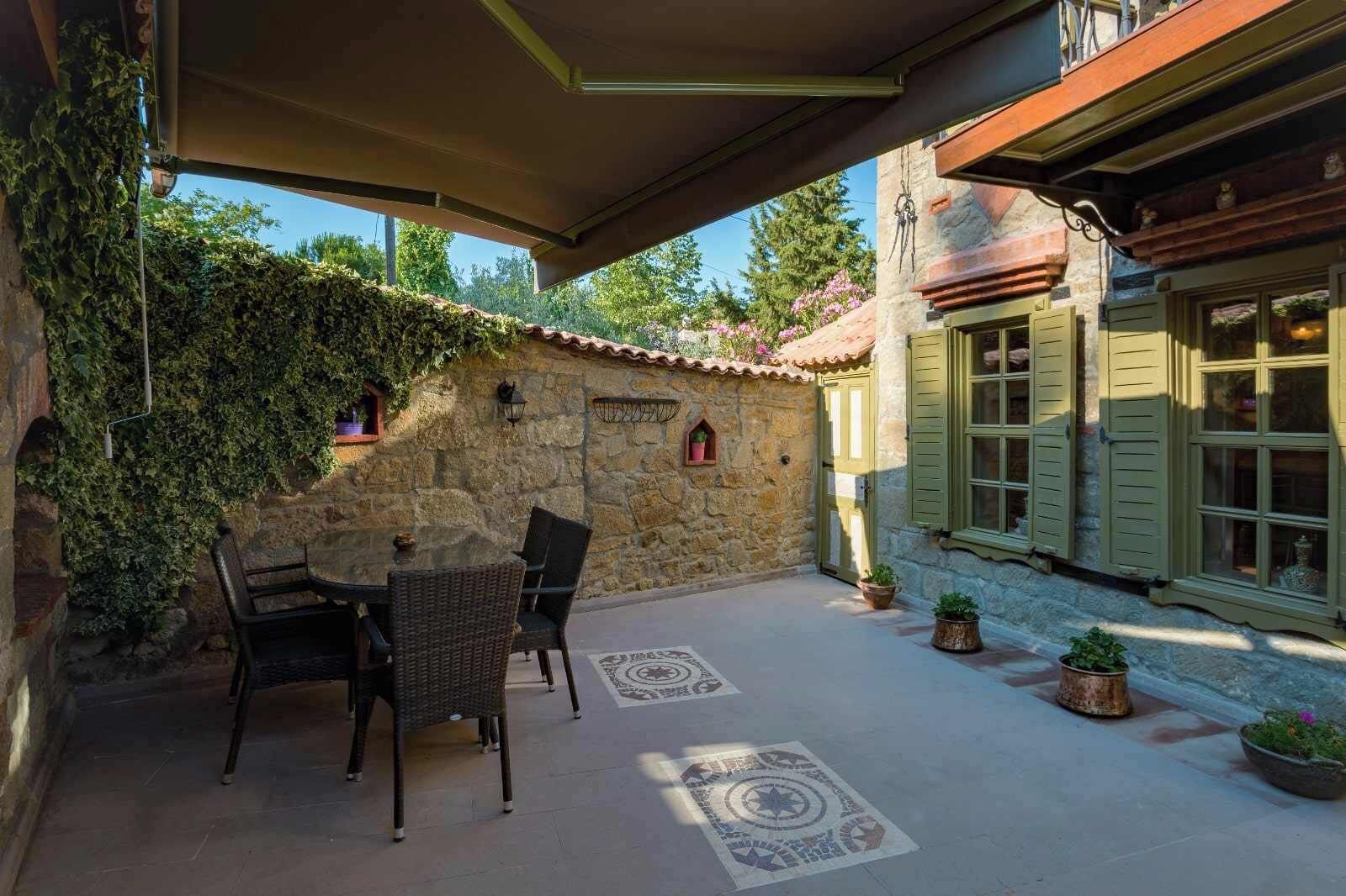A stone patio with a table and chairs, a covered area, and a building with shutters and a tiled roof.