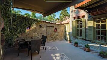 A stone patio with a table and chairs, a covered area, and a building with shutters and a tiled roof.