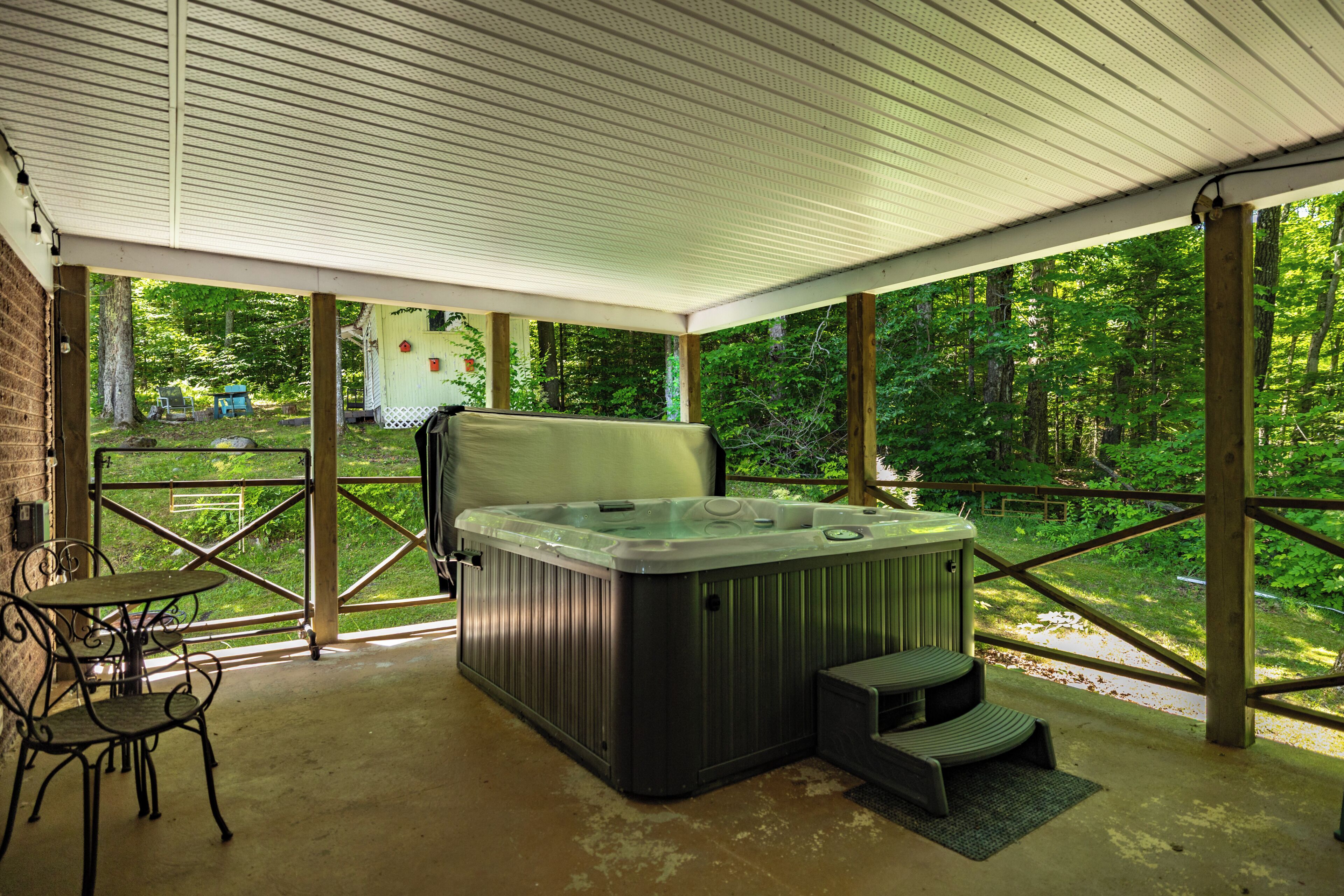 A covered outdoor area with a hot tub, seating, and a view of a wooded area.