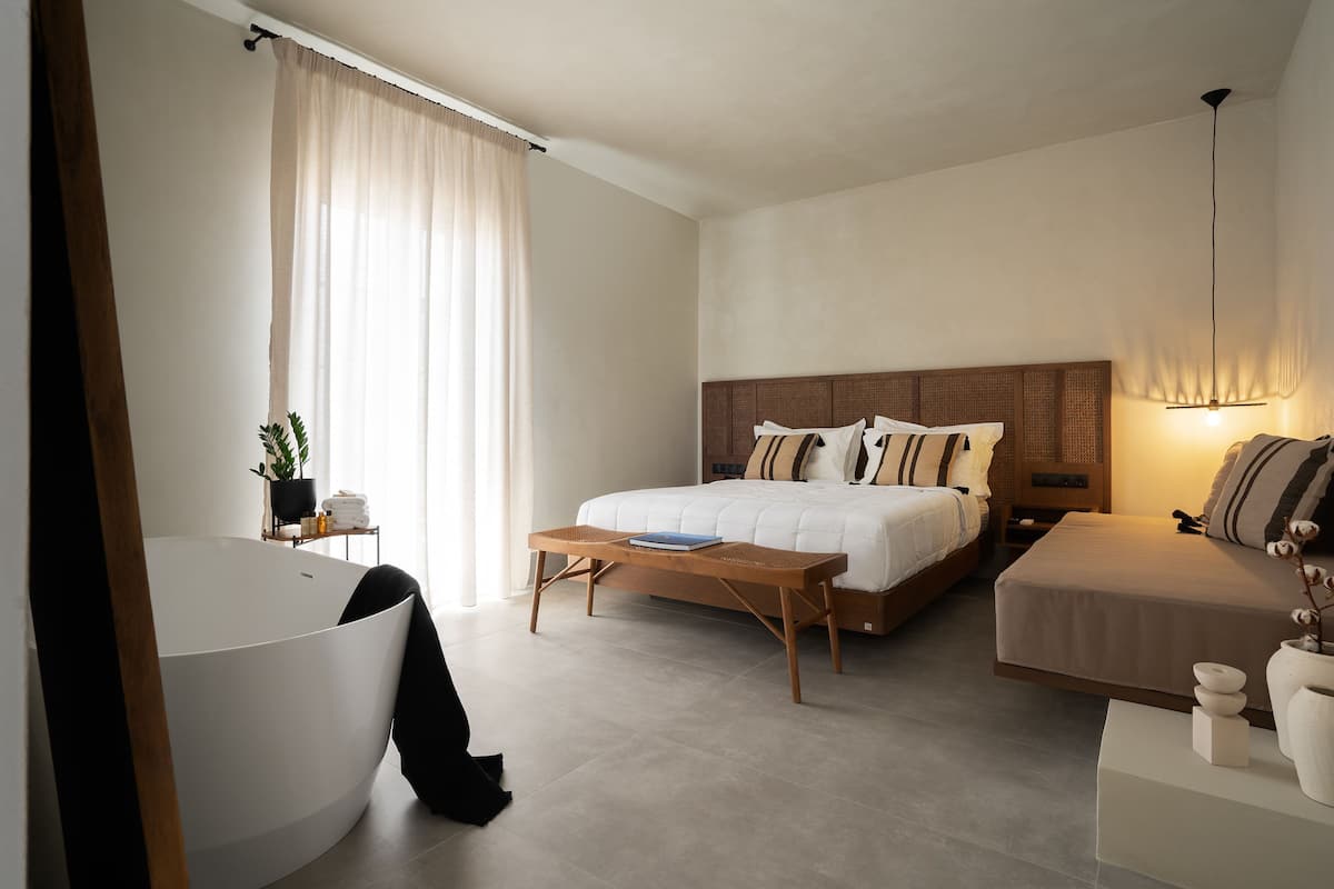 Een moderne hotelkamer met een groot bad, een bed met een houten hoofdeinde, een nachtkastje met een lamp en een kleine plant.
