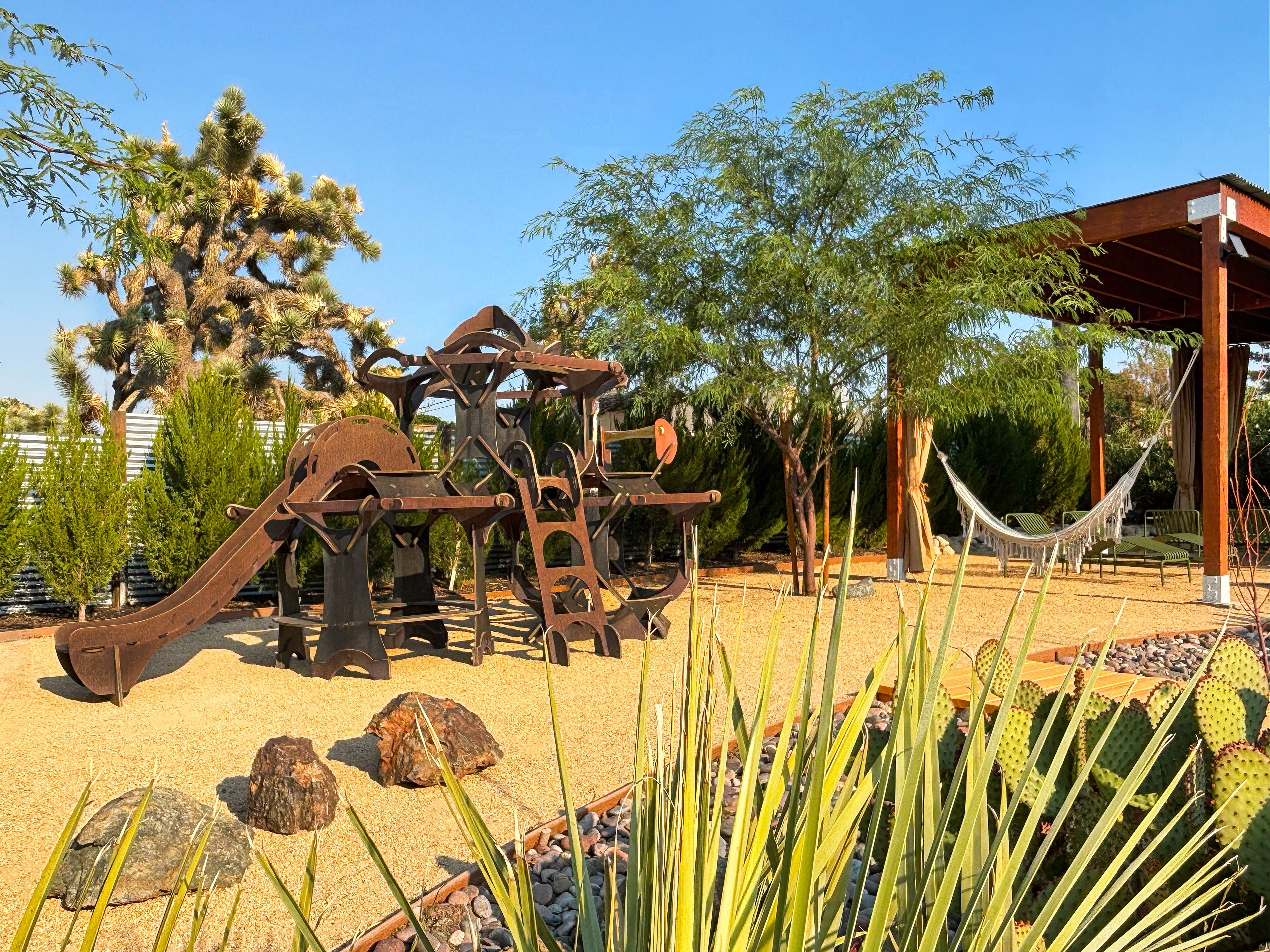 Un parc infantil amb tobogan i elements per escalar, envoltat de cactus i arbres.