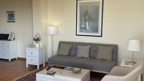 Un salon avec un canapé gris, une table basse blanche, un tapis à motifs, un lustre et une œuvre d’art encadrée accrochée au mur.