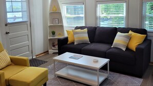 Un salón con sofá negro, cojines amarillos, una mesa de centro blanca y un sillón amarillo. Hay tres lámparas colgantes sobre el sofá.