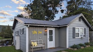Una pequeña casa de un solo piso con fachada gris y una puerta blanca. En el exterior, hay un banco con cojines amarillos y un cartel en la pared que dice 'Lilly Pad'.