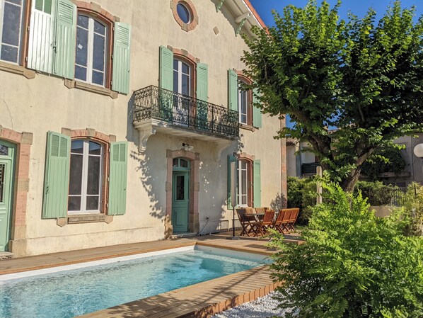 A two-story building with a swimming pool, green shutters, and a balcony with a wrought iron railing.