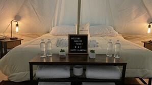 Eine gemütliche Zelteinrichtung mit Bett, zwei Nachttischlampen, einem Willkommensschild, Wasserflaschen, einer kleinen Pflanze und ordentlich gefalteten Handtüchern.
