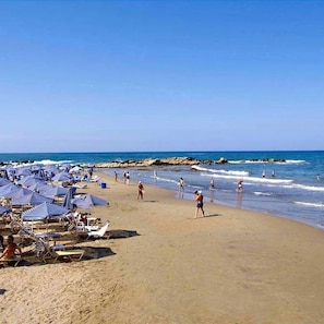 Una playa con muchos sombrillas y tumbonas, orilla arenosa y el océano al fondo.