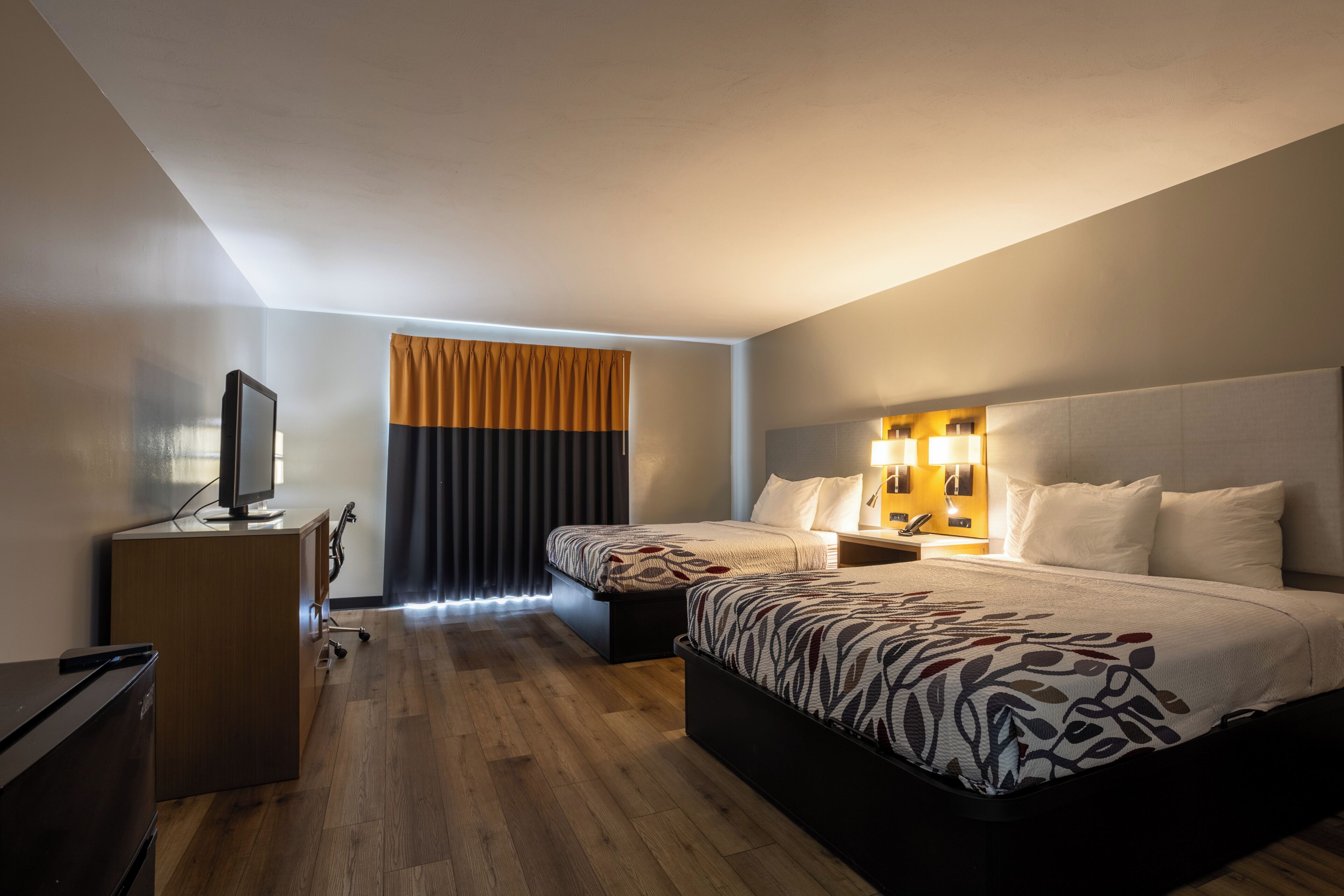 A hotel room with a bed, bedside tables, a desk, a television, and a window with curtains.