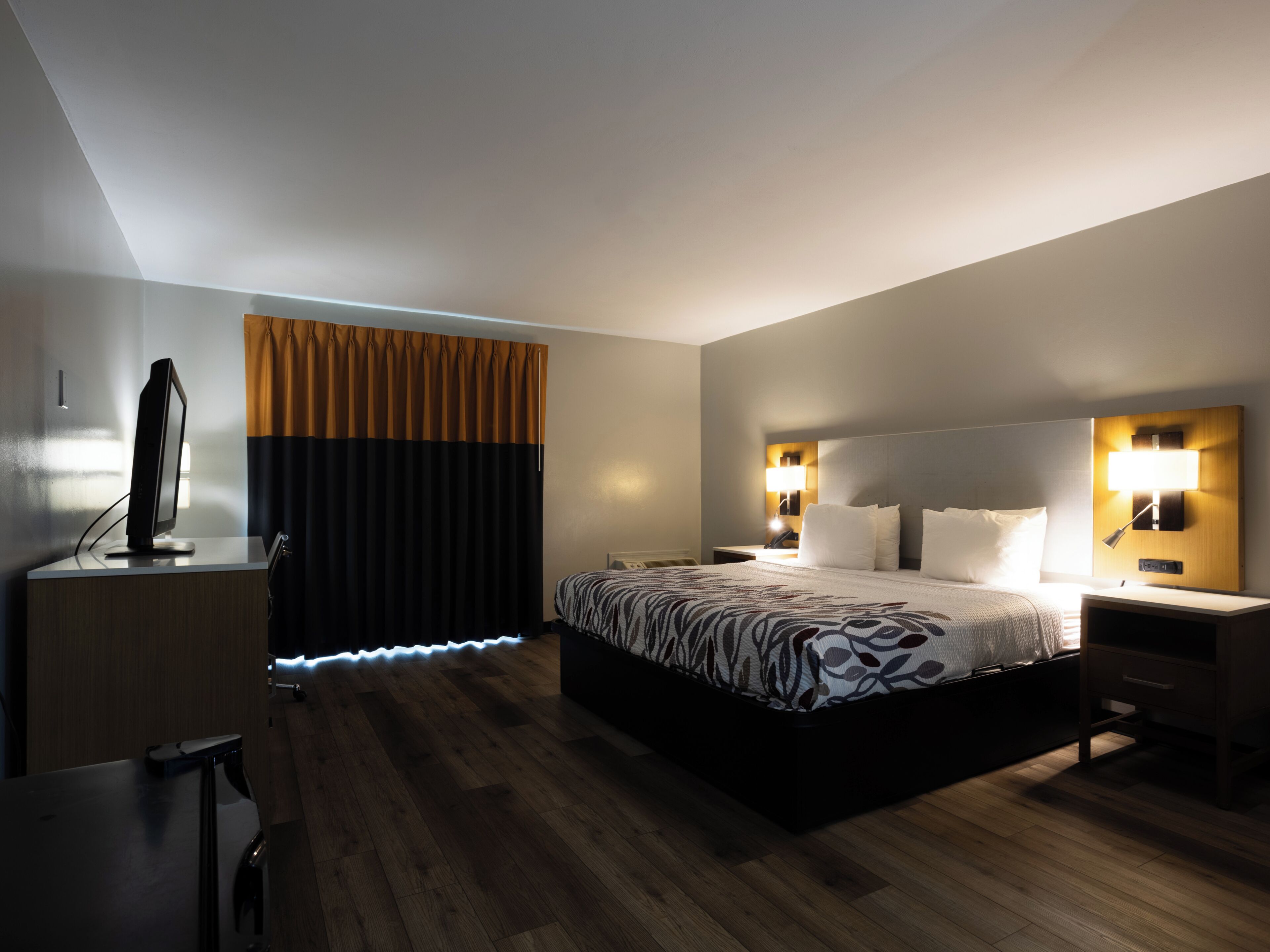 A hotel room with a large bed, bedside lamps, a desk with a computer, and a window with curtains.