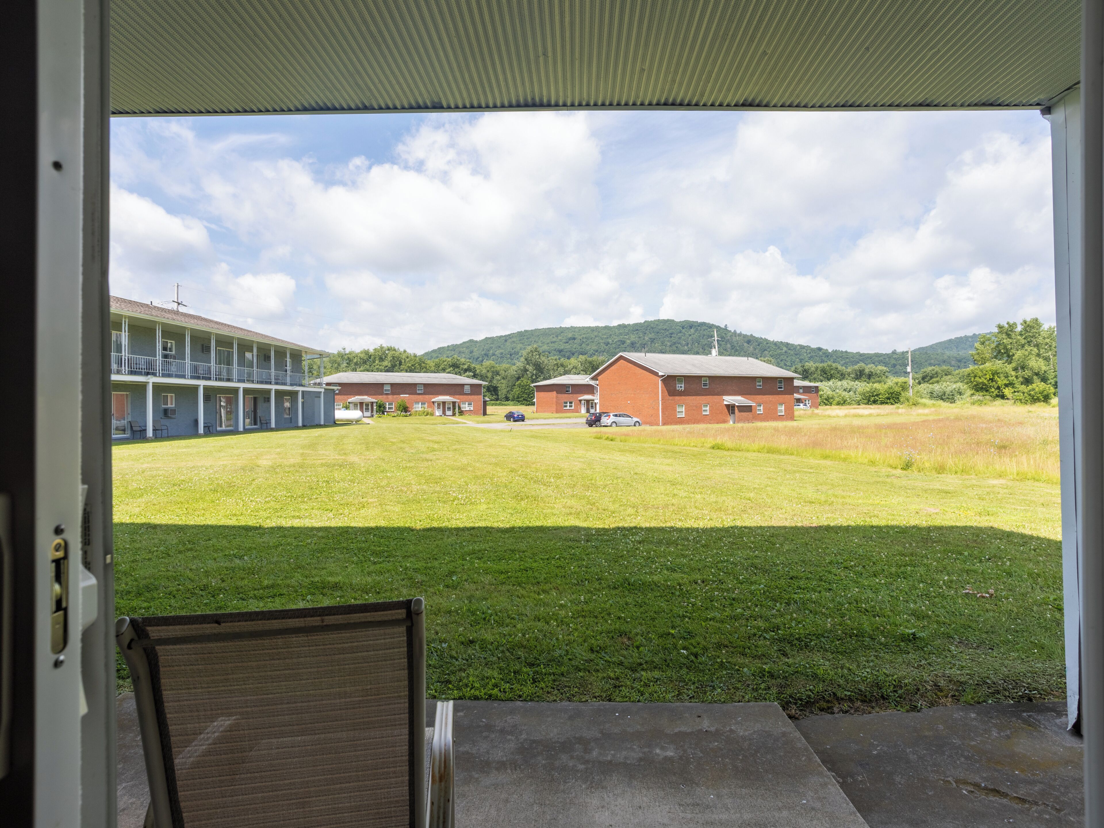 A view from a room with a large window overlooking a grassy field, a building with a red brick facade, and a blue building with a balcony.