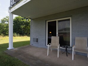 Un patio cubierto con dos sillas y una pequeña mesa, una puerta corrediza de vidrio que da a una habitación interior, y una pared exterior de ladrillo.