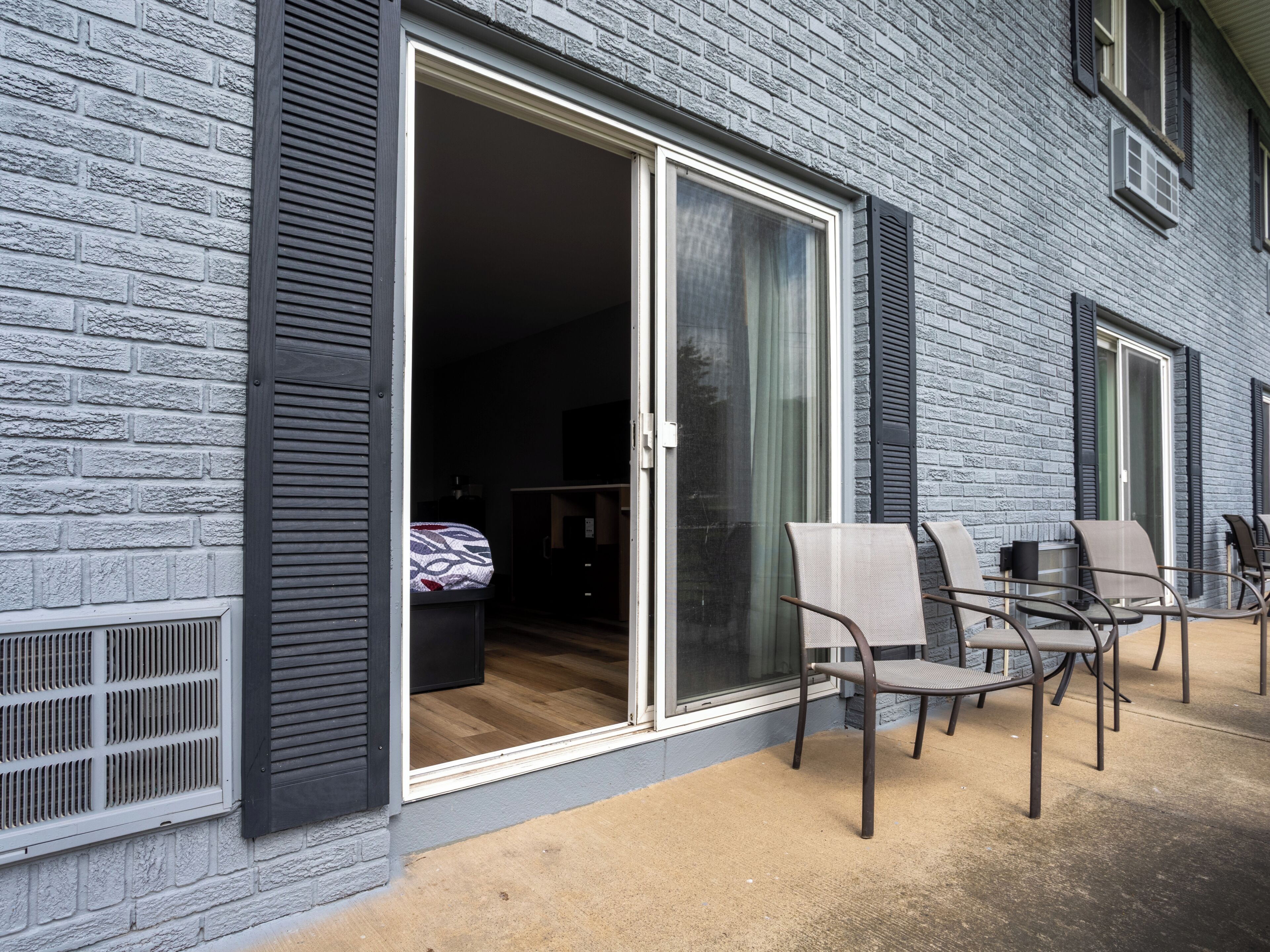 A building with a brick exterior, a sliding glass door leading to a room with a bed and a desk, and outdoor seating with chairs.