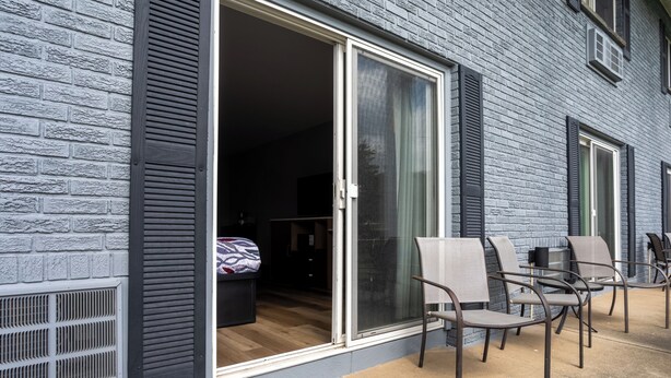 A building with a brick exterior, a sliding glass door leading to a room with a bed and a desk, and outdoor seating with chairs.