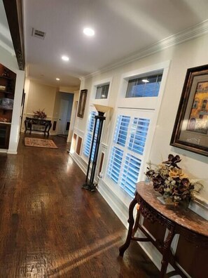 A hallway with wooden flooring, a decorative table with flowers, and a window with blinds.