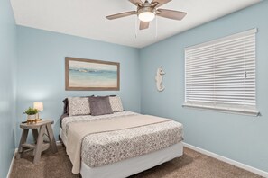 Une chambre à coucher avec un lit, un ventilateur de plafond, une fenêtre avec des stores, une table de chevet avec une lampe et un tableau encadré au mur.