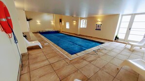 An indoor swimming pool with tiled floors, white poolside chairs, and wall-mounted lights. - Former showhome with indoor pool & gym (Oakhill)