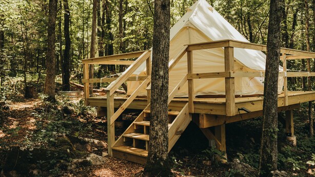 Ein hölzernes Baumhaus mit einem Dreiecks-Dach und einer Holztreppe, die zu ihm hinauf führt, umgeben von einem Wald.