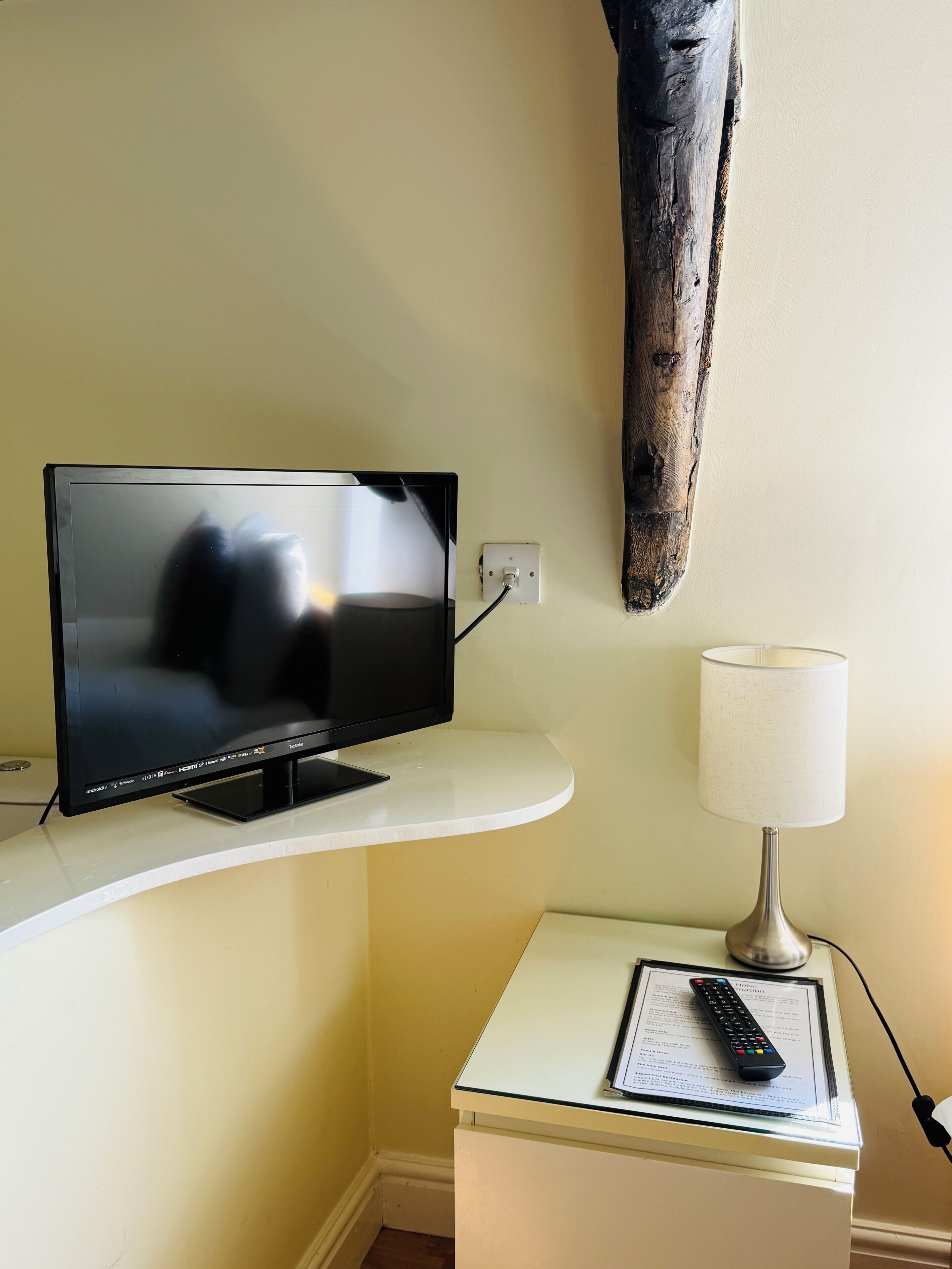 Un televisor sobre un pedestal blanco, un control remoto sobre una mesa de vidrio y una lámpara sobre una mesita.