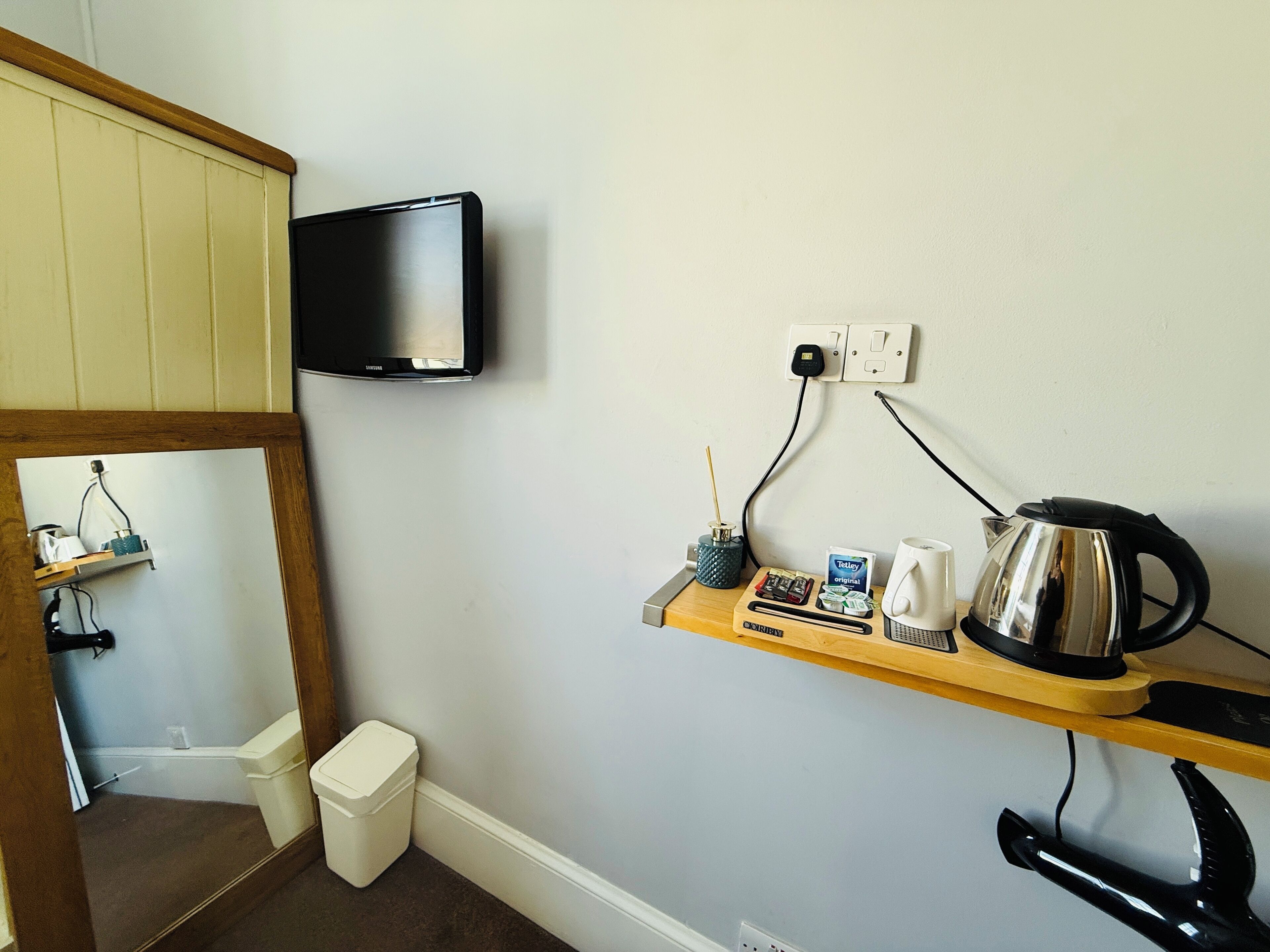 A small room with a wooden shelf holding a kettle, a cup, a box of tissues, and a small plant. A television is mounted on the wall above the shelf.
